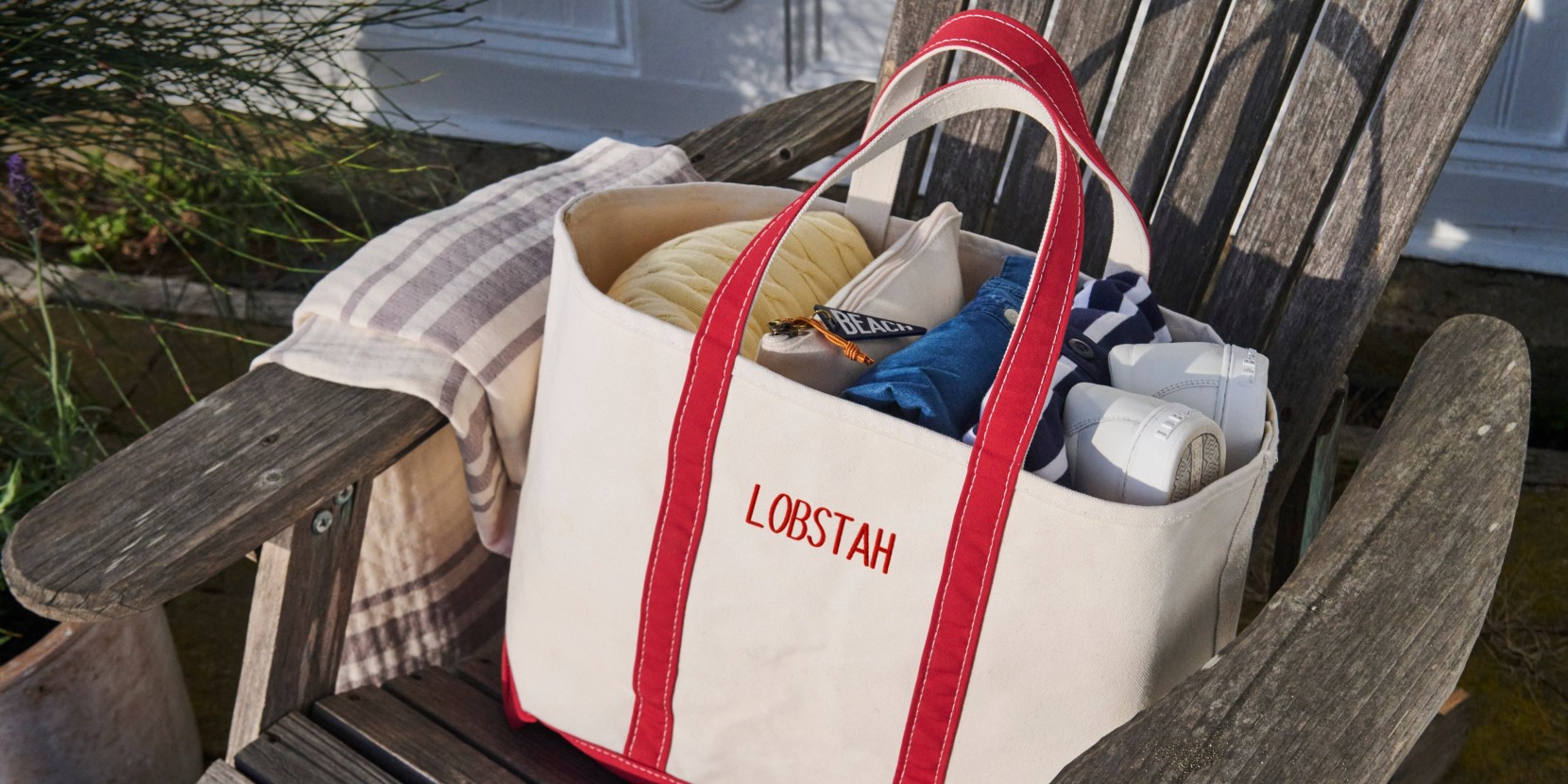 Canvas tote with red handles reading “LOBSTAH” sits on wooden bench, filled with towels, clothes, and shoes.