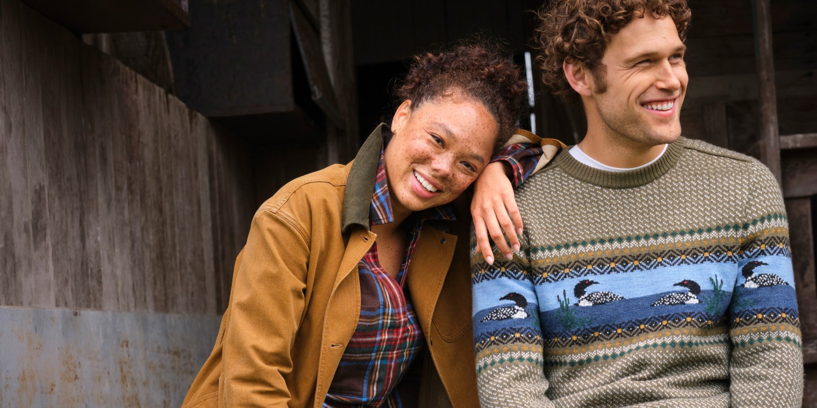 Two people in fall layers sit outside a rustic building; one wears a loon-pattern sweater and the other a plaid shirt and jacket.