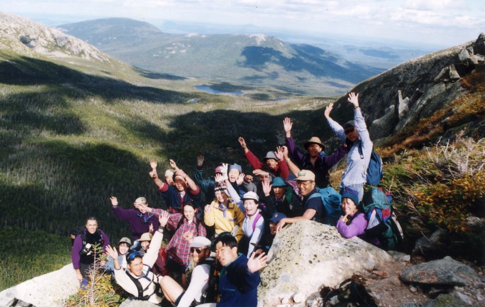 Vintage photo of a large group of people waving while on a hiking trail with a vast mountainous landscape behind them.