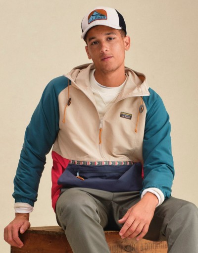 Person wearing a colorblock anorak with teal sleeves and tan front, paired with a logo cap and casual pants.