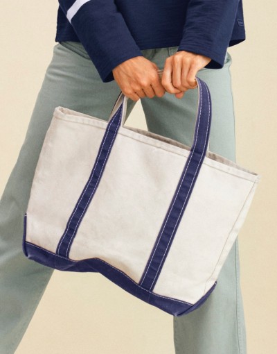 Close up image of a canvas tote bag with navy trim held by a person wearing light pants and a dark long-sleeve top.