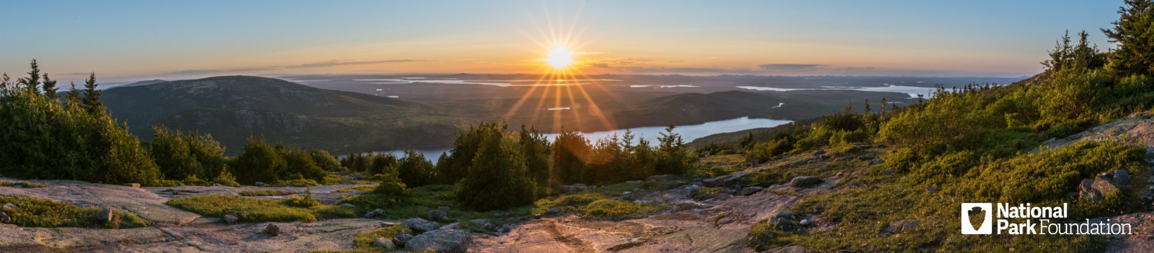 Sunrise over forested mountains and winding lakes, seen from a rocky trail, with National Park Foundation logo.