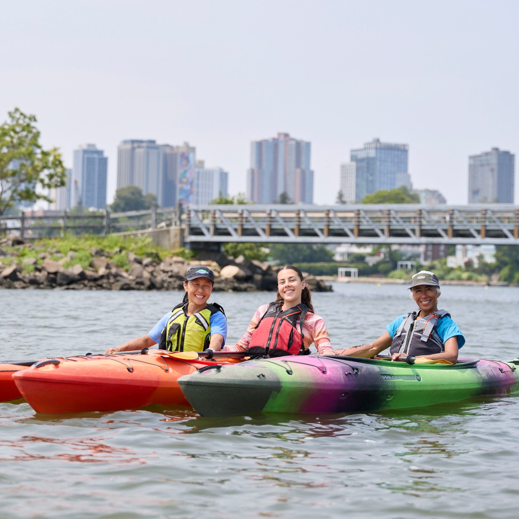 3 smiling people in kayaks on the water, a city skyline in the distance.