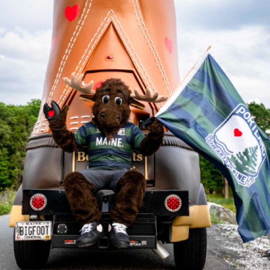 Someone in a moose costume waving a Maine Hearts of Pine flag while sitting on the rear bumper of the Boot mobile.