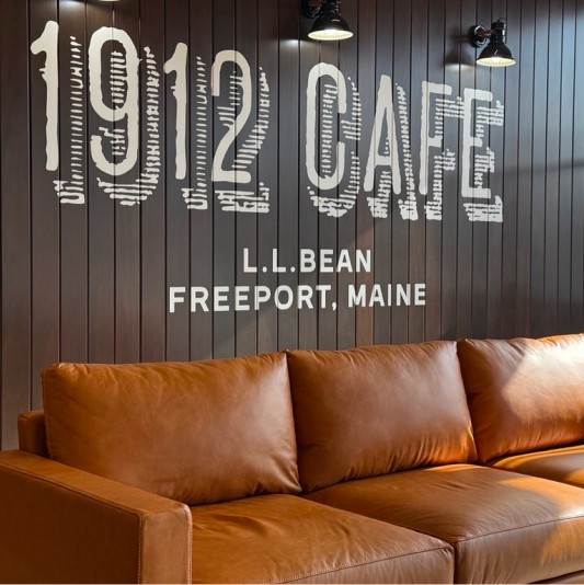 Deep brown slat wall with cream-colored "1912 Cafe L.L.Bean Freeport, Maine" painted on it, an inviting leather couch below.