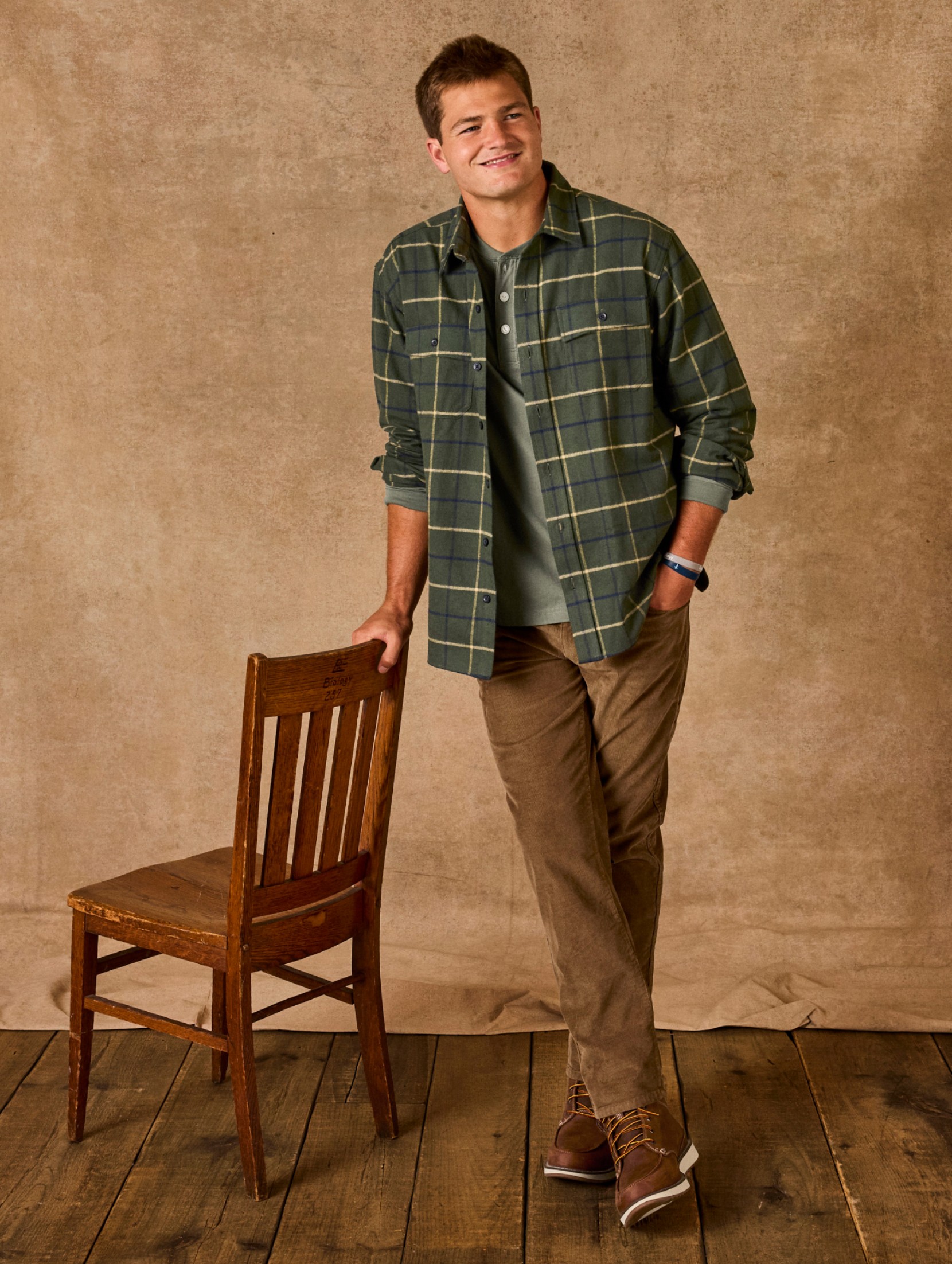 Drake Maye standing, leaning on a wooden chair, wearing Beanflex Cord 5-Pocket Pants and a plaid chamois shirt.