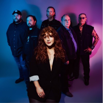 Lexi James standing in front of 5 band members, in blue and purple lit space.