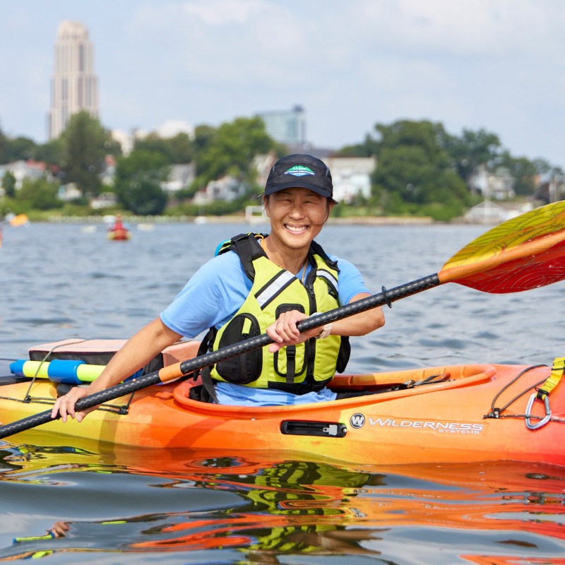 Person wearing a life vest and paddling a colorful kayak on a river, with trees and city buildings in the background.