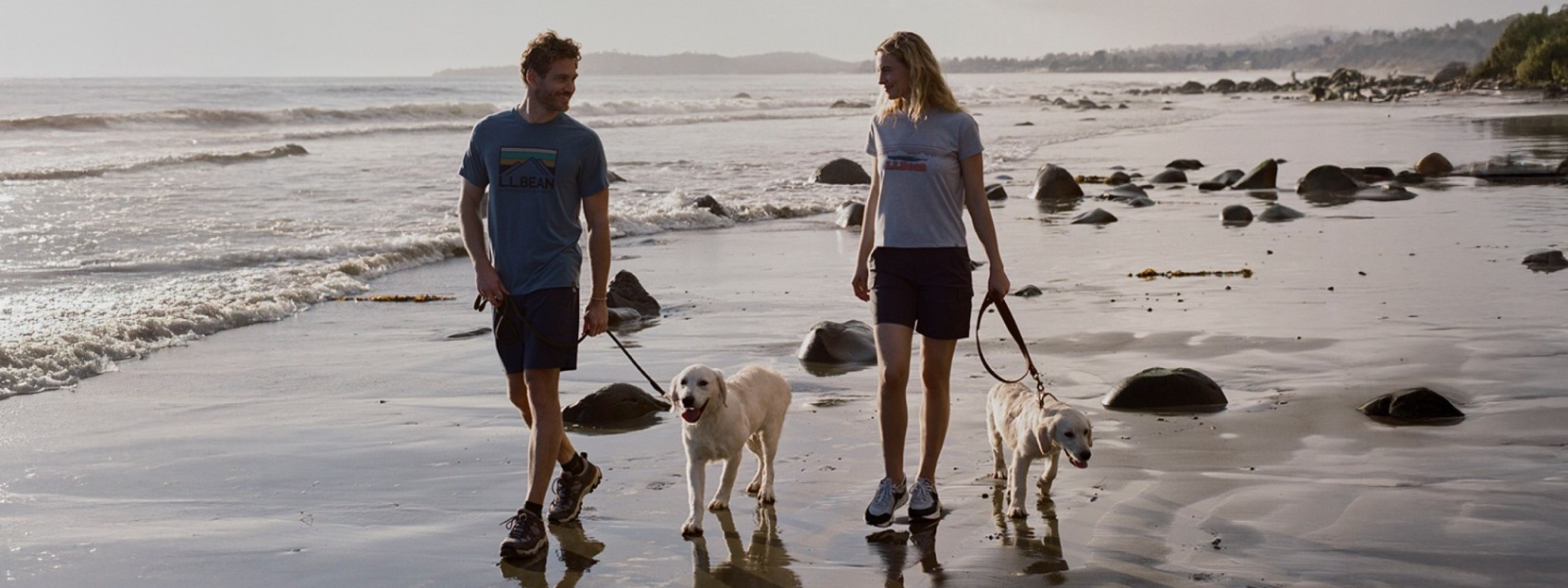 Two people in summer clothes walk dogs along a rocky beach shoreline with waves gently rolling in under soft light.