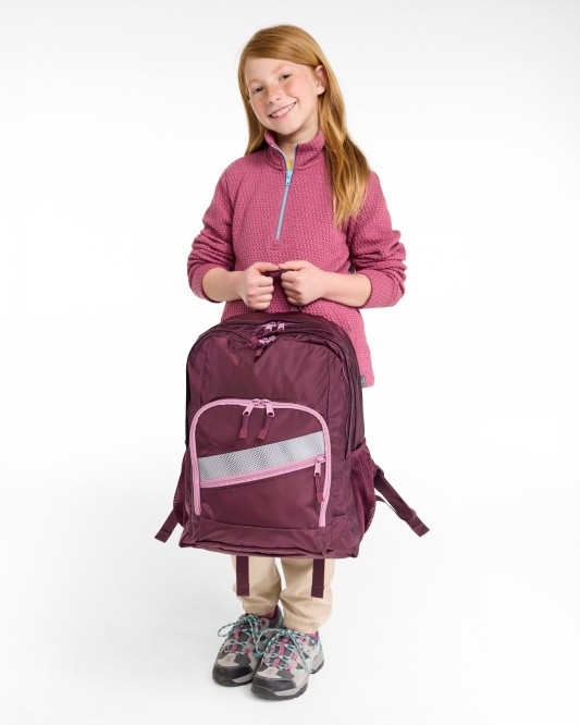 Smiling child holds a maroon backpack, wearing a pink pullover, beige pants, and sneakers against a plain background.