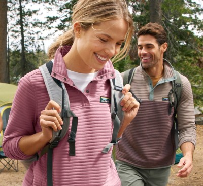 Two smiling people wearing L. L. Bean colorblock pullovers and backpacks walk through a forested campsite.