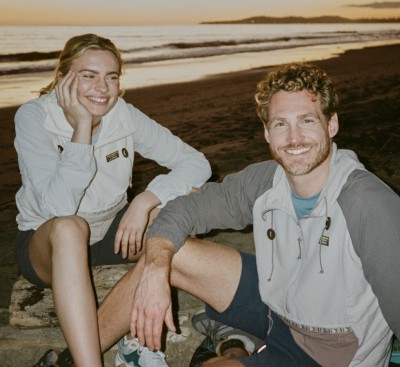 Two people sit on a rocky beach at sunset, wearing light jackets and shorts, with ocean waves and distant hills behind.