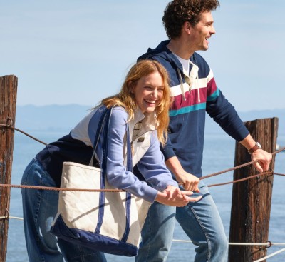 Two people stand on a waterfront pier, leaning on rope rails with calm water and distant hills behind them.