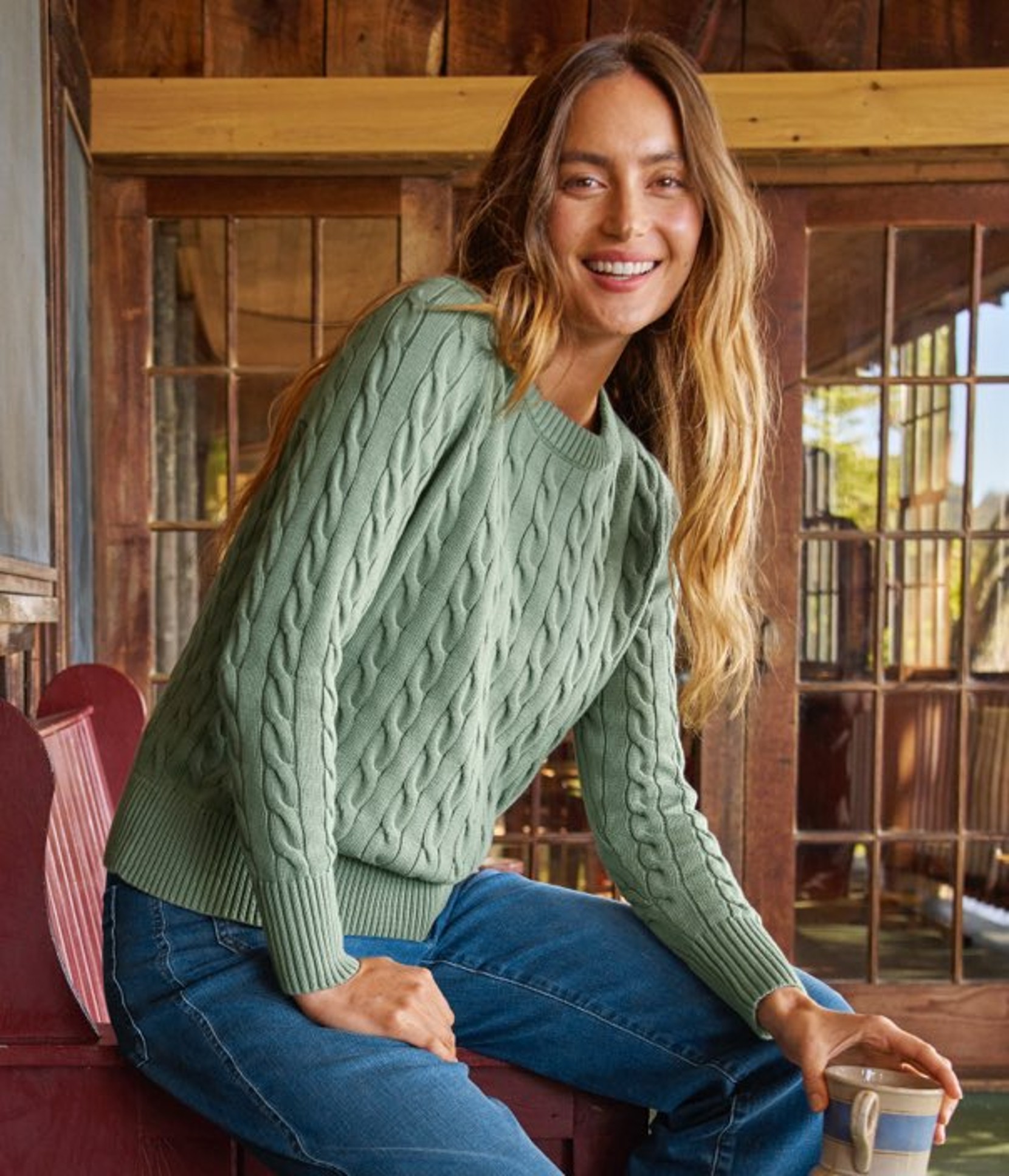Woman wearing a mint cable‑knit sweater and blue jeans, seated indoors in a rustic wooden setting.
