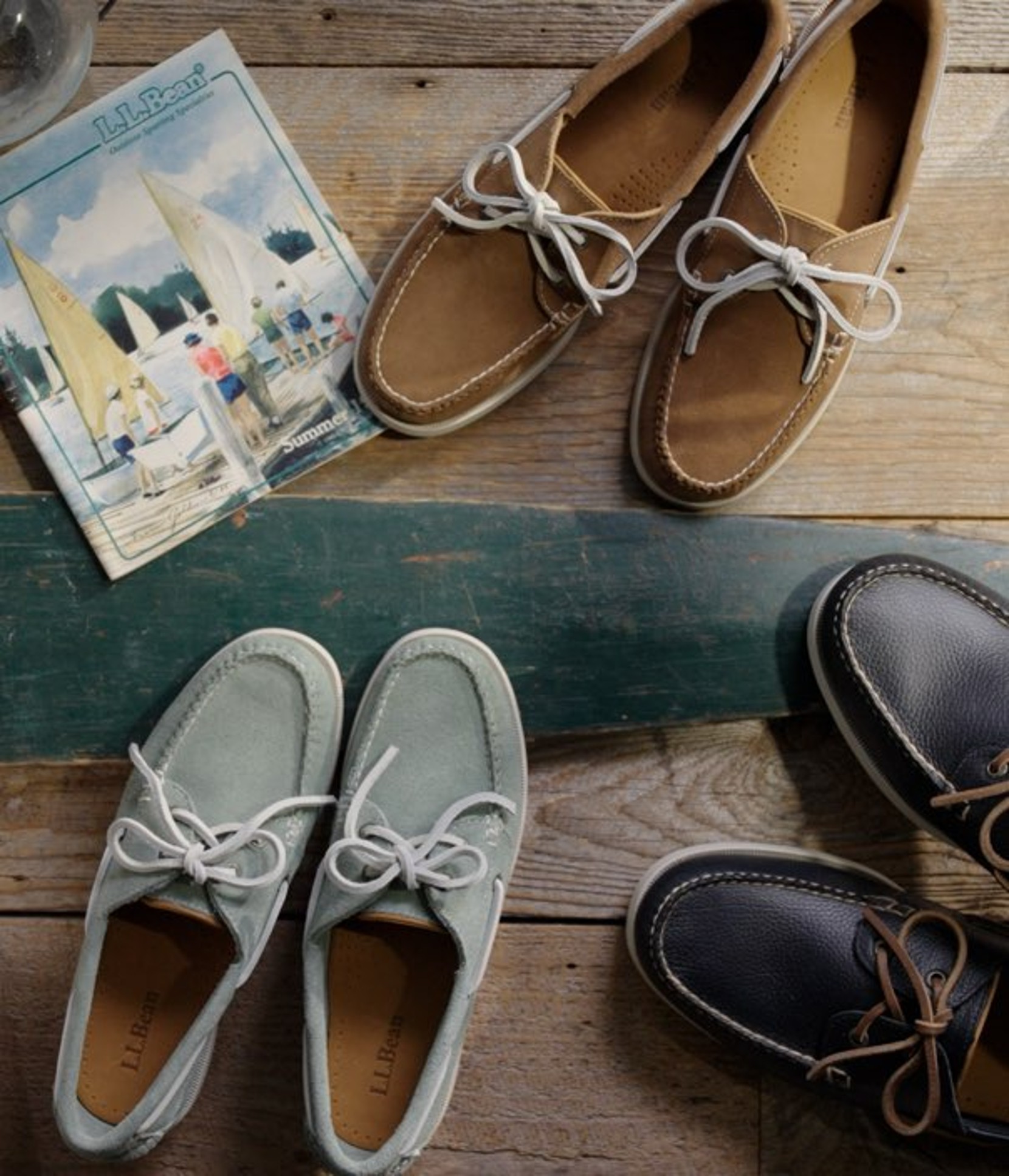 Vintage nautical-themed display with several pairs of boat shoes, an oar, and a catalog on wooden planks.