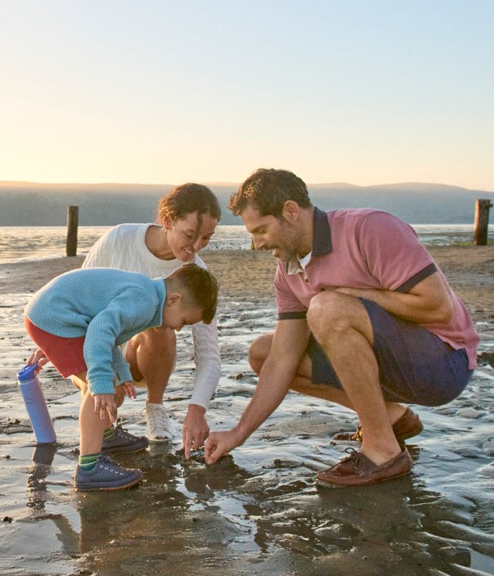 Three people crouch on a sandy beach, examining tide pools at sunset with a calm sea in the background.