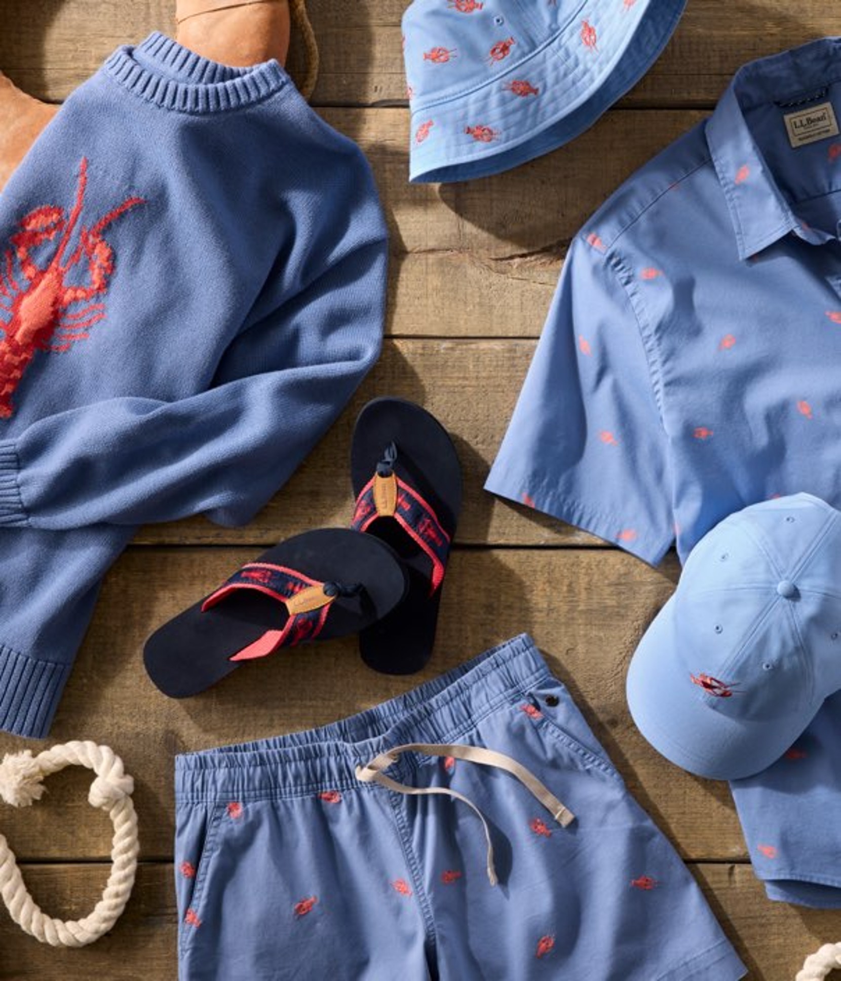 Flat lay of blue lobster-print sweater, shirt, shorts, hat and flip-flops arranged on a wooden surface.