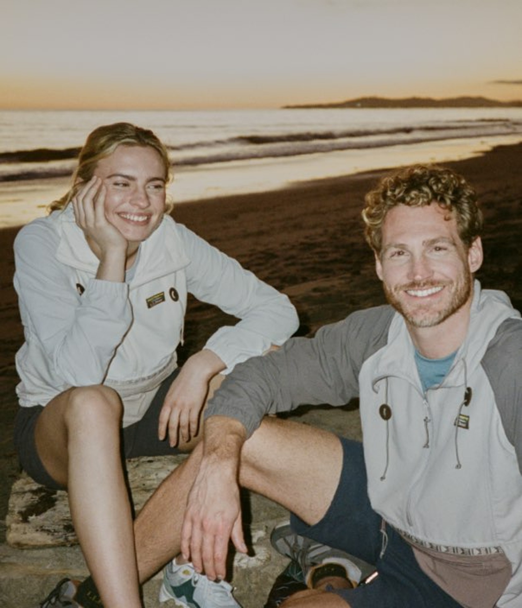 Two people sit on a rocky beach at sunset, wearing light jackets and shorts, with ocean waves and distant hills behind.