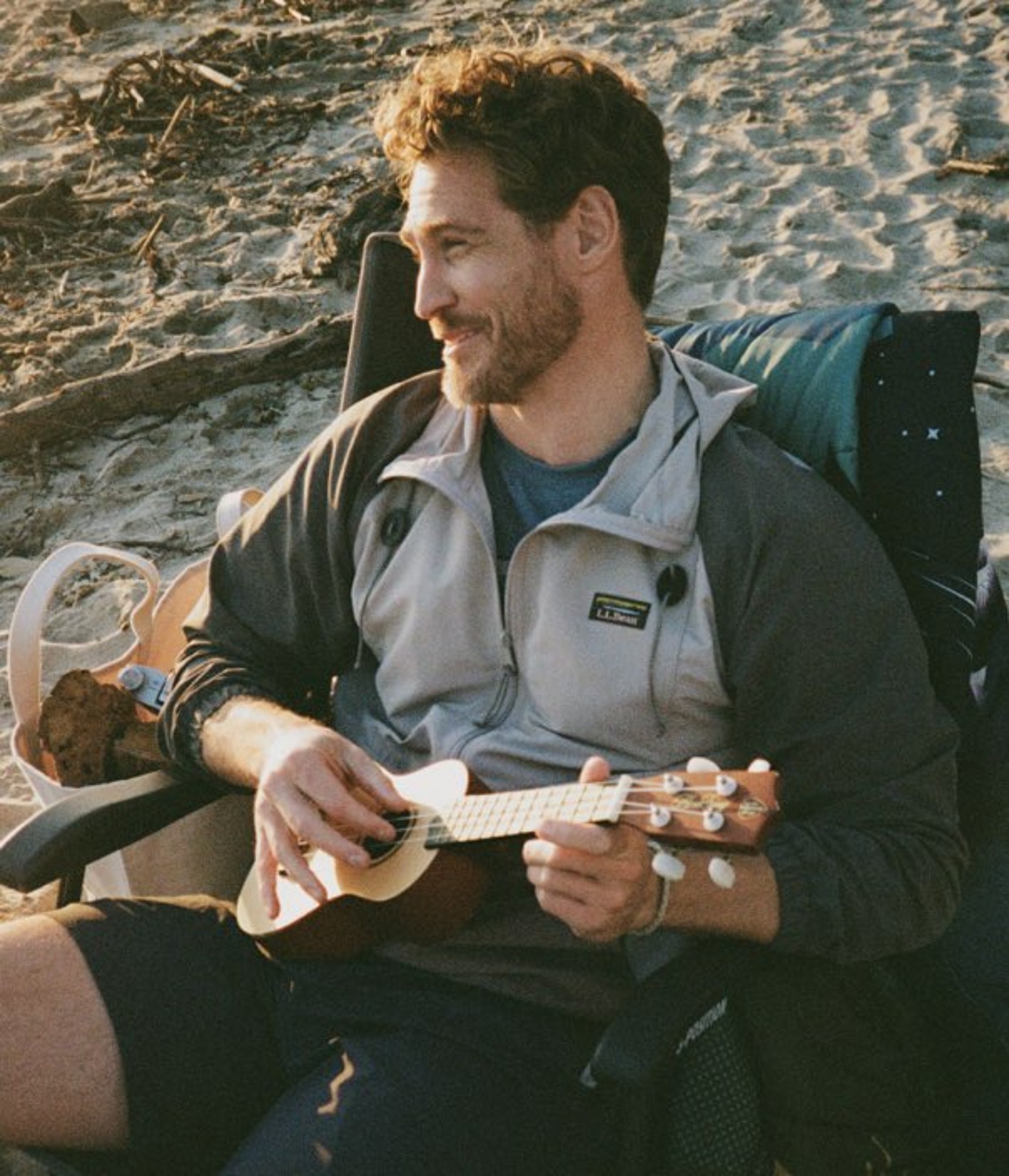 A person sits on a beach chair holding a ukulele, with sand behind, a cooler nearby, and camping gear around.