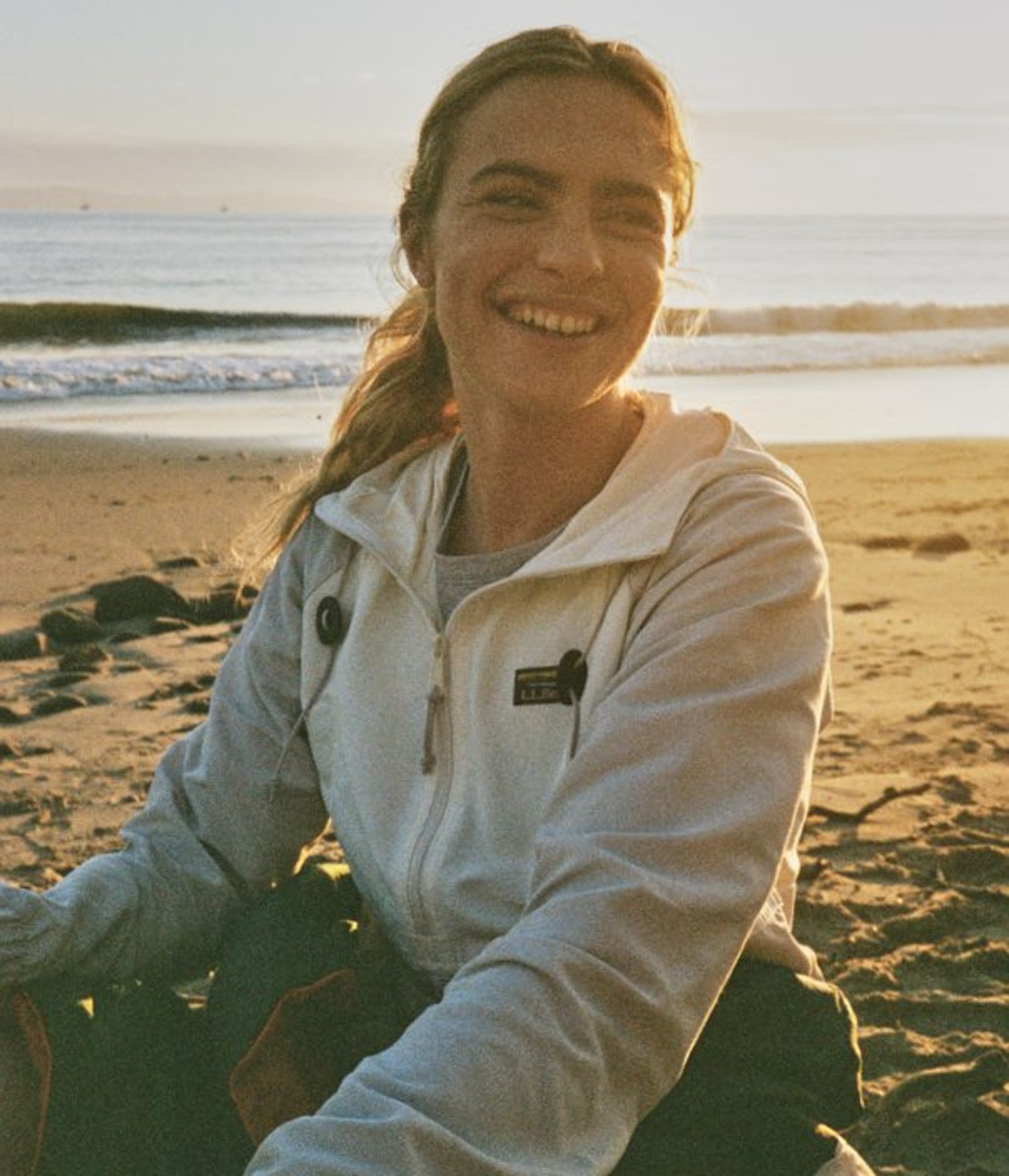 A person sits on a sandy beach at sunset, wearing a light jacket, with ocean waves behind.