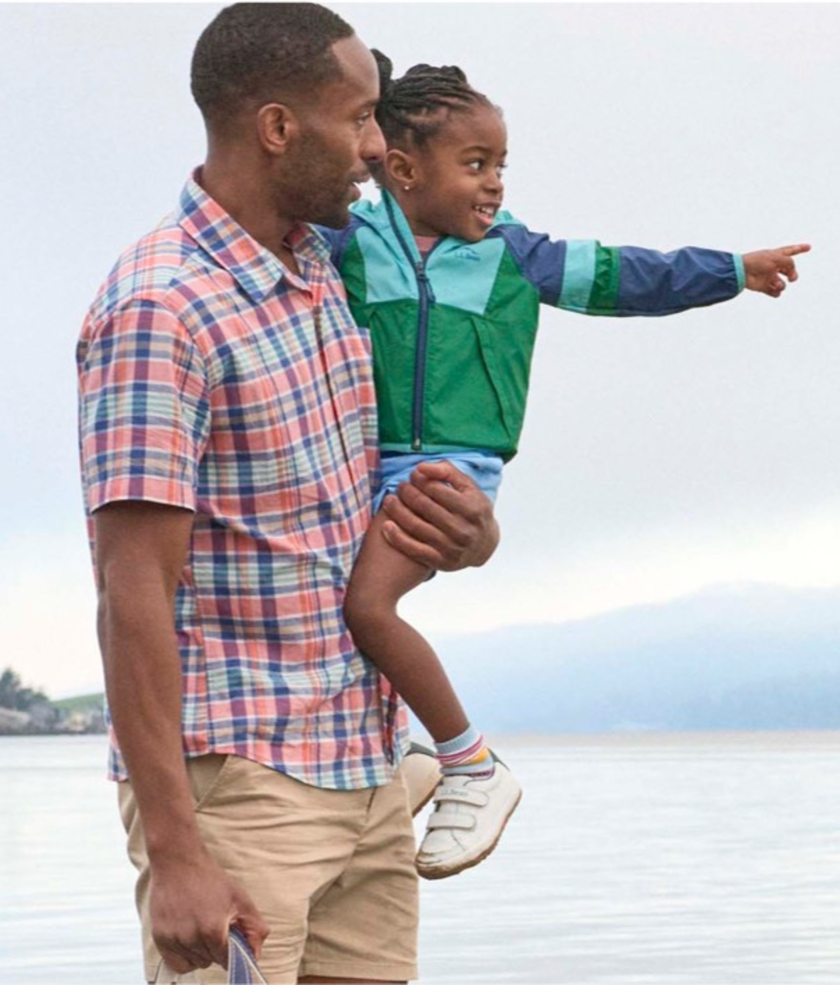 Adult carrying a child by the water as the child points into the distance; cloudy sky and shoreline in background.