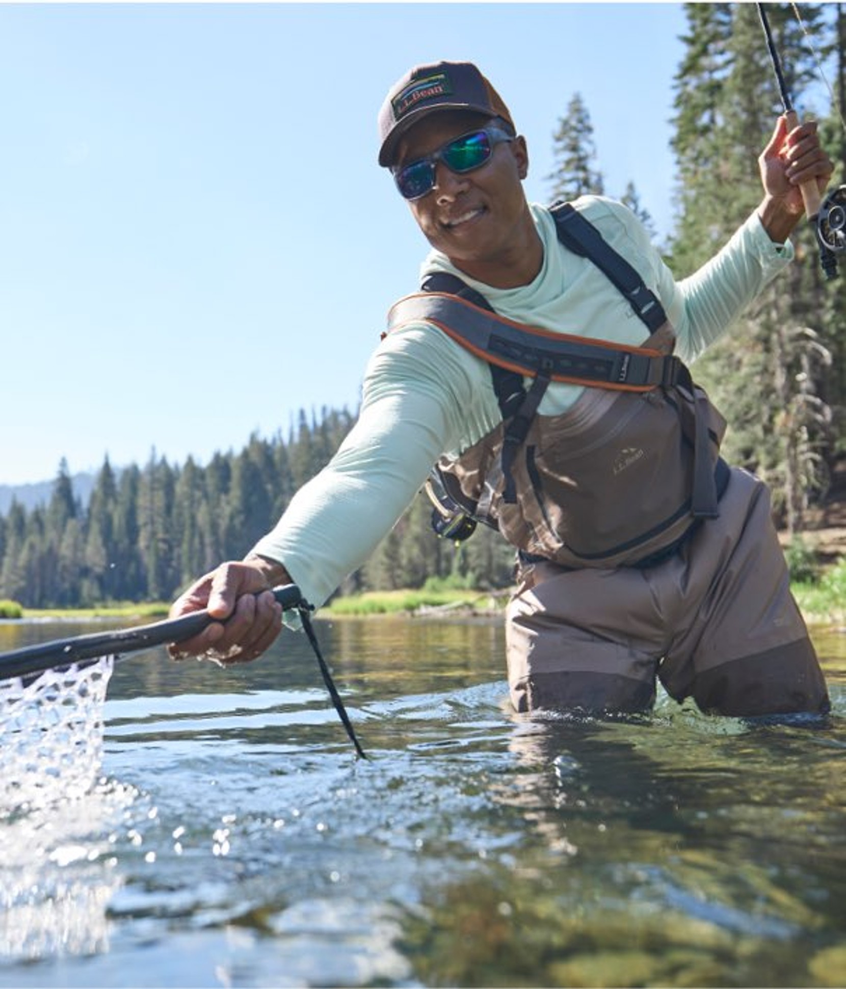 Person in waders stands in a river, using a net while fly fishing near a forested shoreline on a clear day.