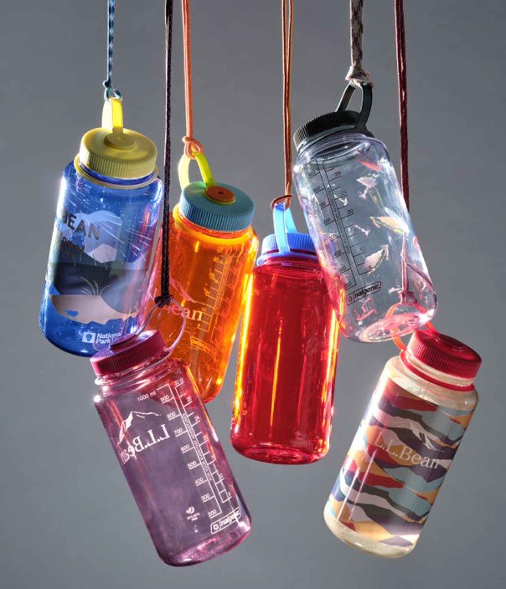 Six colorful reusable water bottles hang by cords against a gray background, glowing as light shines through translucent plastic.