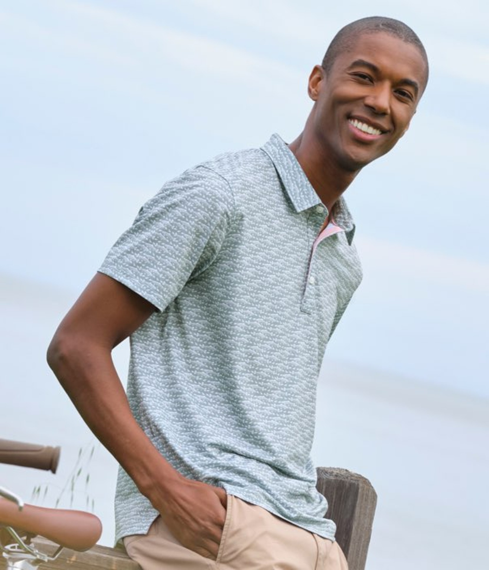 Person in a light patterned polo and khaki pants stands beside a bicycle and wooden post under a cloudy sky.