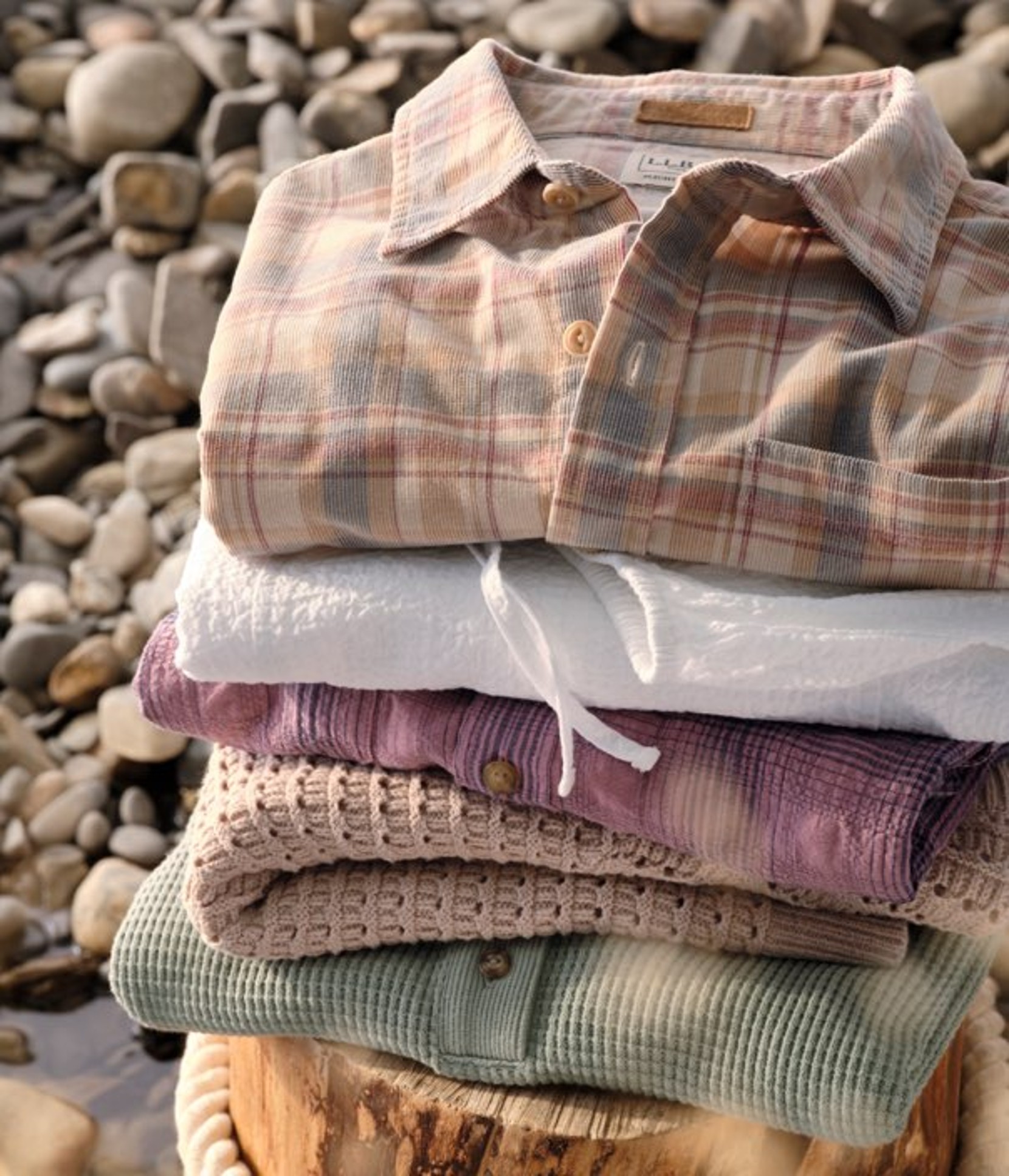 Stack of folded pastel shirts placed on rocks, with soft textures and natural tones in an outdoor setting.