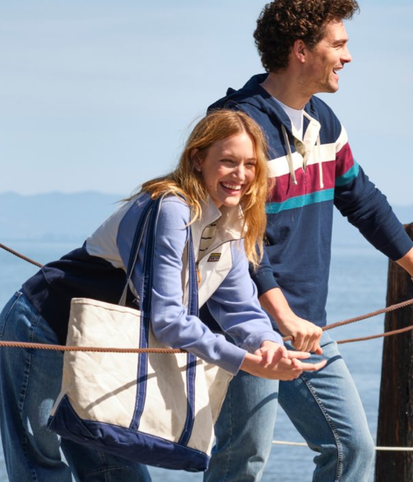 Two people stand on a waterfront pier, leaning on rope rails with calm water and distant hills behind them.