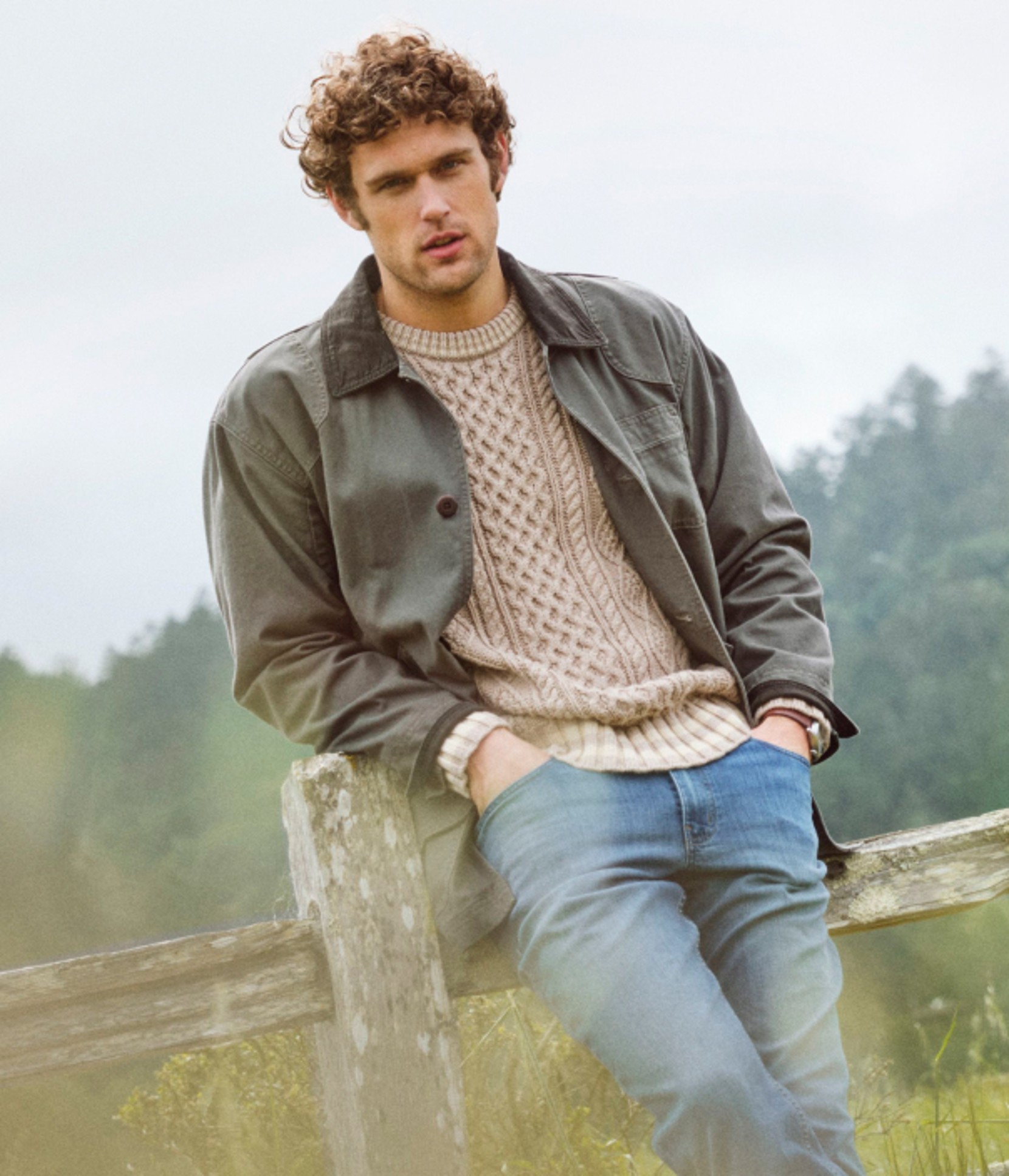 Person leaning on a rustic wooden fence outdoors wearing a jacket over a white sweater.
