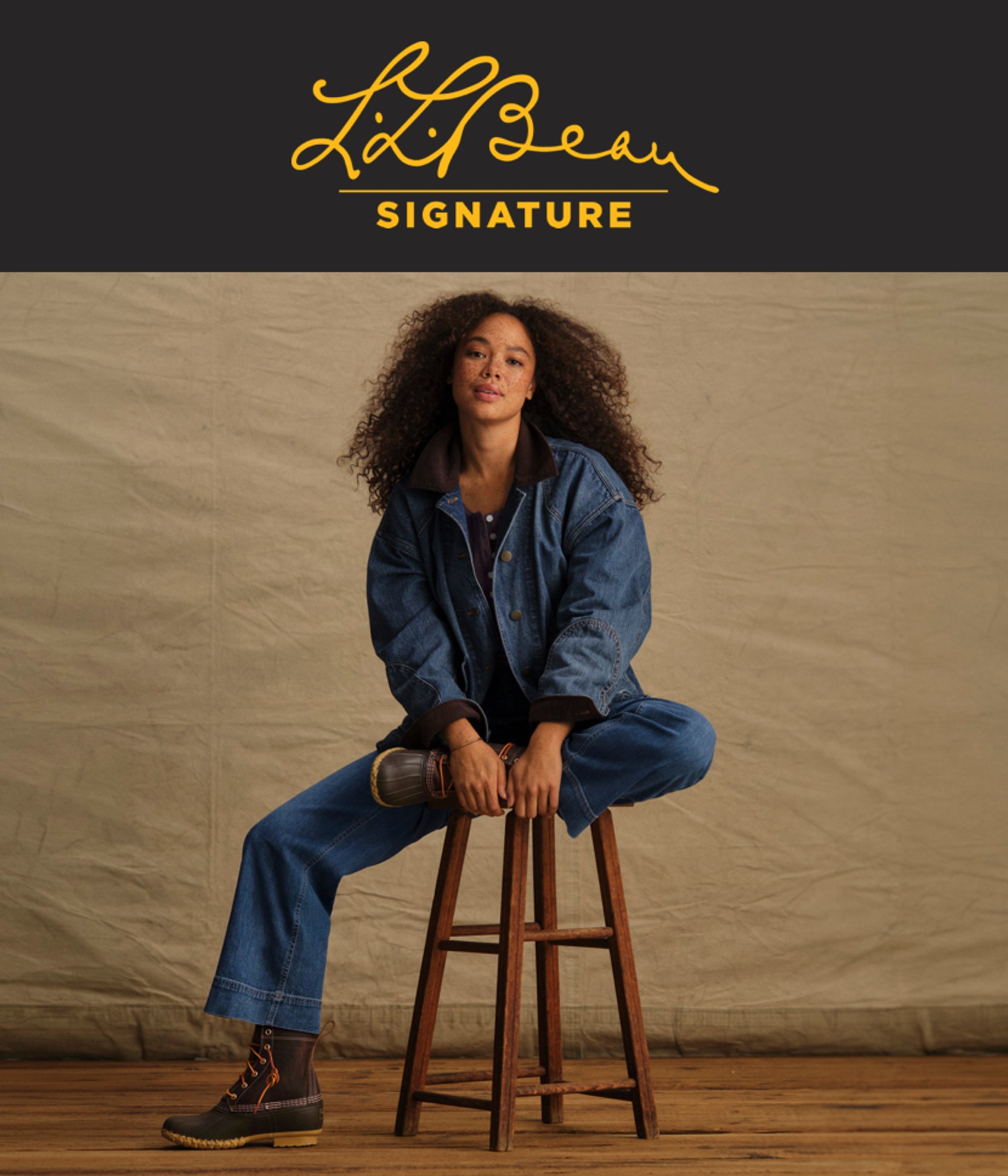 Woman seated on a wooden stool wearing a denim jacket, jeans, and lace-up boots against a neutral backdrop.