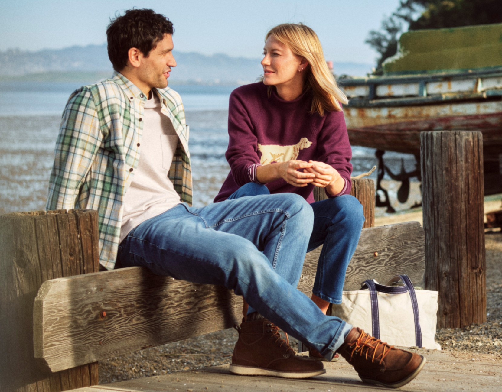 Two people in casual fall clothing sit on a wooden dock by the water, with a canvas tote bag and boat in the background.