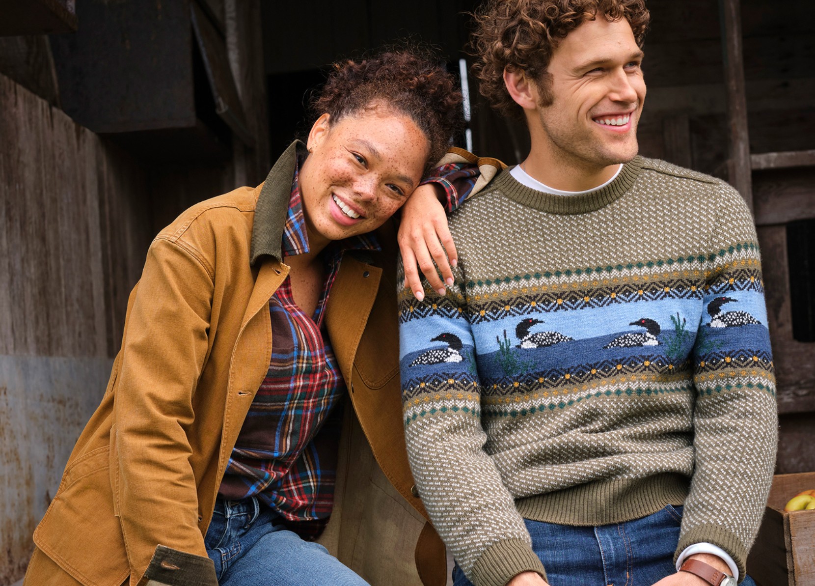 Two people in fall layers sit outside a rustic building; one wears a loon-pattern sweater and the other a plaid shirt and jacket.