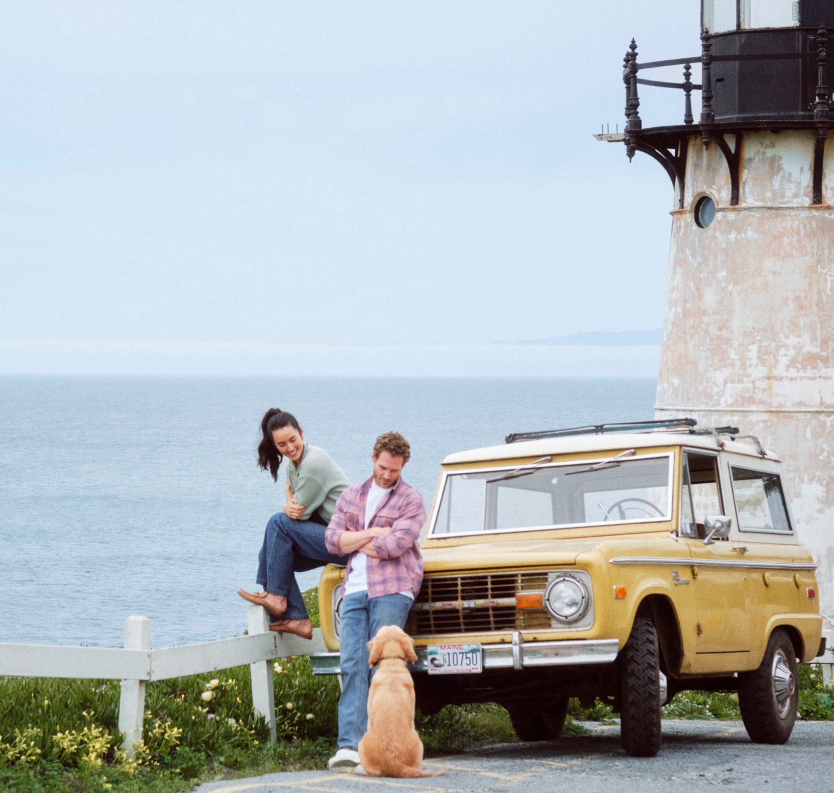 Two people sit by a coastal fence beside a yellow vintage SUV, with a dog standing nearby and ocean cliffs in the background.