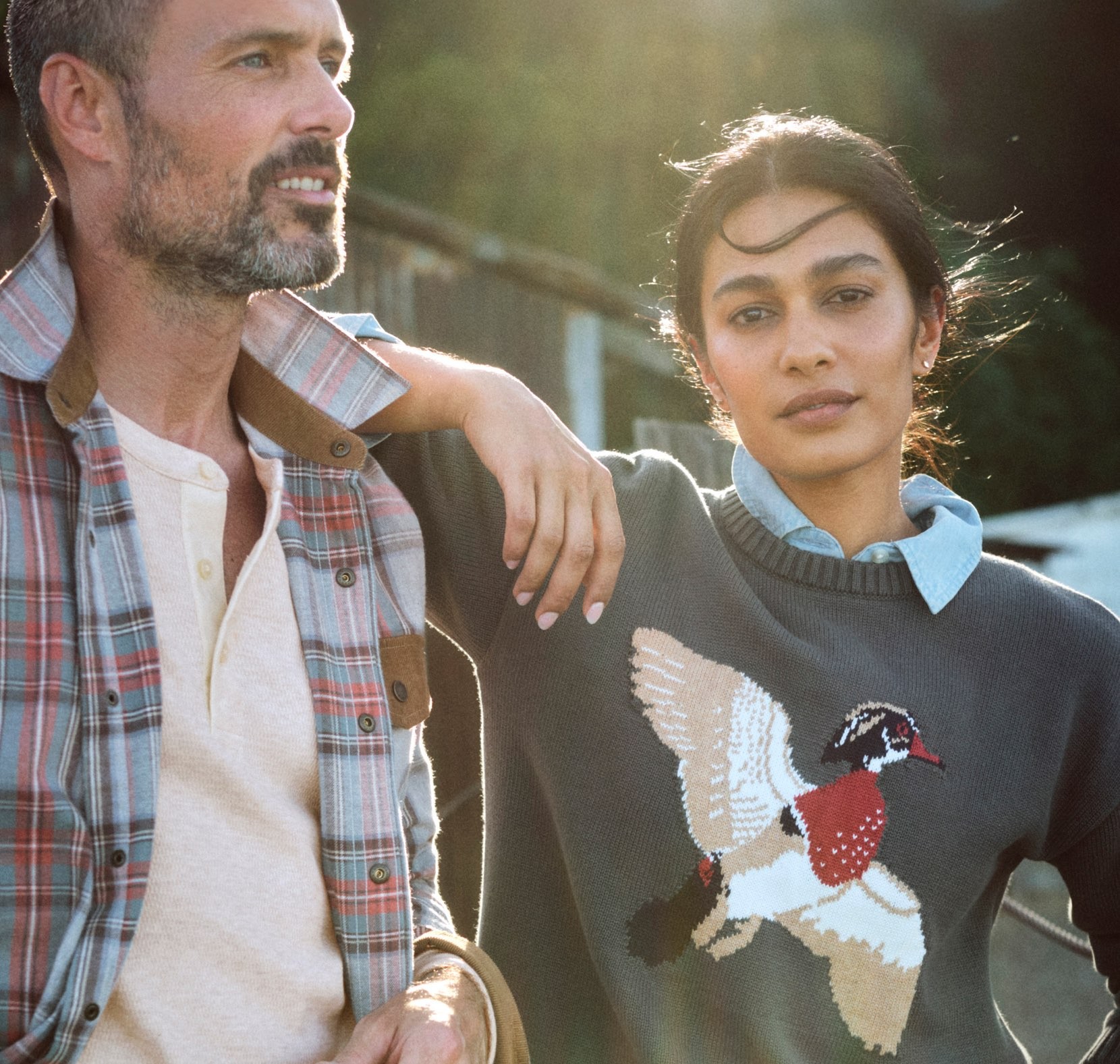 Two people outdoors in warm sunlight, one wearing a bird-patterned sweater and leaning on a companion's shoulder.