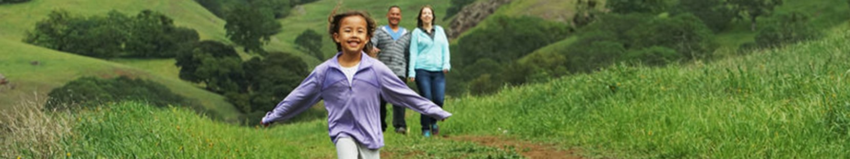 Girl running in a green field with parents walking behind her. 