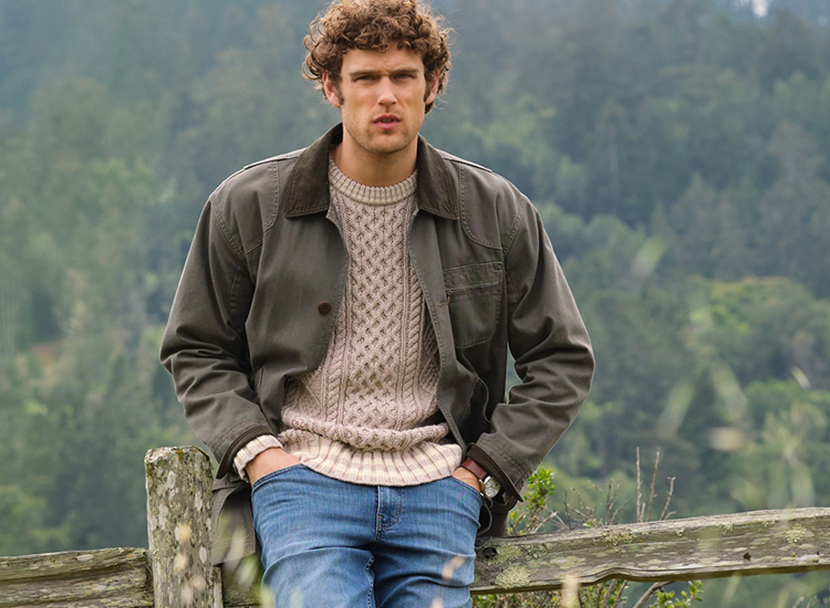 Man wearing a cable-knit sweater, jacket, and jeans sitting on a rustic fence outdoors.