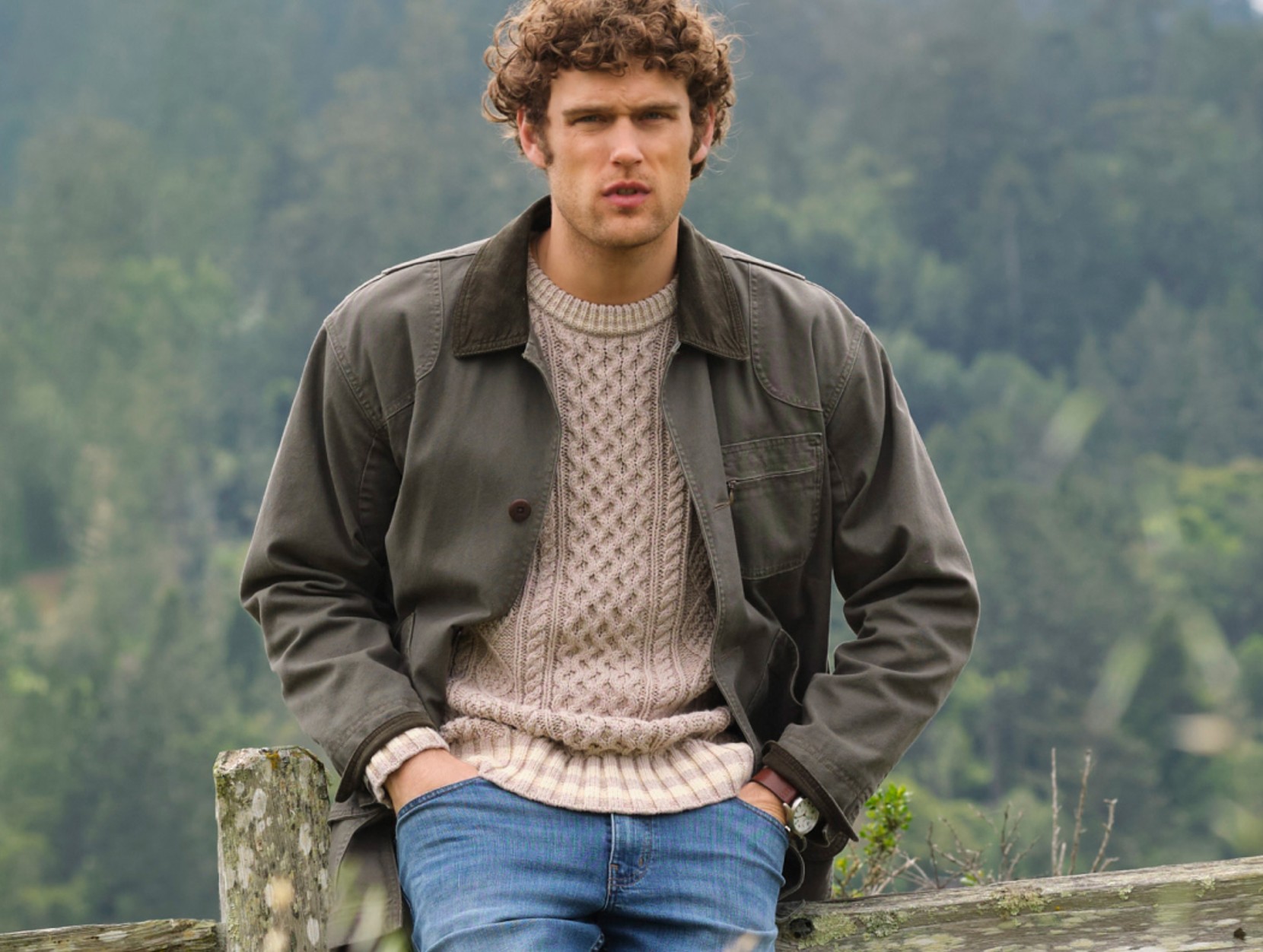 Man wearing a cable-knit sweater, jacket, and jeans sitting on a rustic fence outdoors.