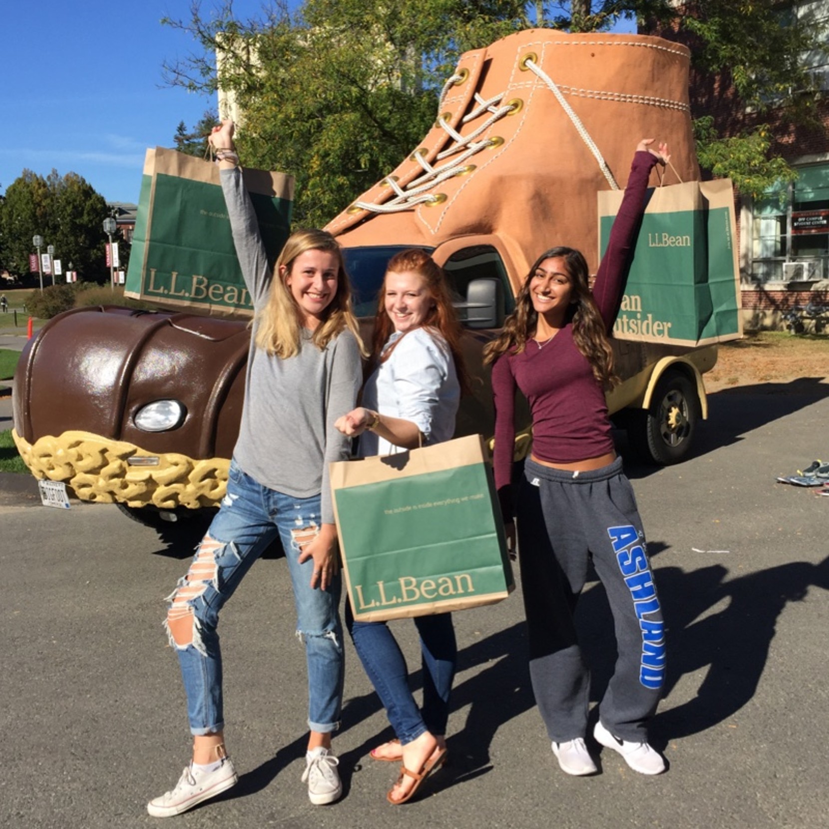 three smiling female college students posing in front of the bootmobile holding shopping bags